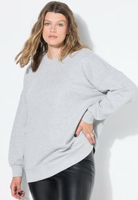 Mladá žena s vlnitými vlasy, která má na sobě kovové náušnice, světle šedou oversized mikinu a černé kožené kalhoty, pózující s jednou rukou na boku.