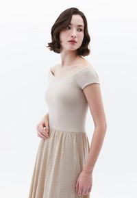 Beige šaty bez ramínek s přiléhavým tělem a strukturovanou, nabíranou sukní. Hladká tkanina nahoře, vzorovaná textura dole.