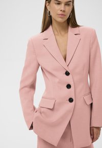 Rosa maßgeschneiderter Blazer mit strukturierter Oberfläche, ausgestattet mit einem Stegkragen, vier schwarzen Knöpfen und zwei Vorderentaschen.