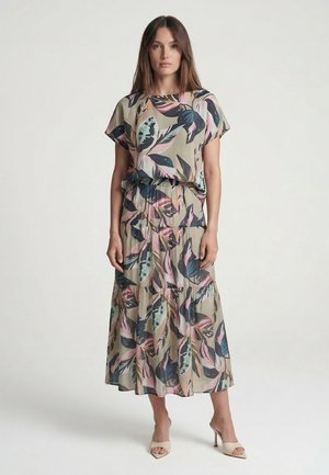 Woman wearing a beige midi dress with colorful large leaf patterns, short sleeves, and a gathered waist, paired with beige heeled sandals.