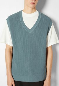 Jasnoniebieski dzianinowy sweter bez rękawów z dekoltem w serek, o fakturowanym wzorze, z prążkowanymi detalami i luźnym krojem noszony na białej koszuli.