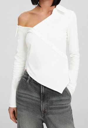 Femme portant un haut asymétrique blanc à épaule dénudée et manches longues associé à un jean noir délavé taille haute, main dans la poche.
