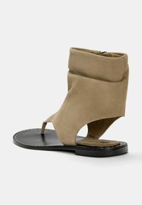 Bež usnjeni sandal z zloženim zgornjim delom, odprtimi prsti in ravnim črnim podplatom. Ima šive ob podplatu in stransko zadrgo.