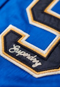 Tissu bleu avec un grand numéro "5" brodé en noir et blanc, bordé de coutures dorées et blanches, avec le texte "Superdry".