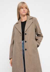 Manteau en mélange de laine brun avec un col cranté, cinq boutons noirs, deux poches latérales et une coupe droite, porté sur un haut noir et un jean bleu.