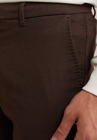 Pantaloni din bumbac maro cu o textură fină, având un buzunar lateral și detalii de cusătură vizibile. Mână plasată pe țesătură.