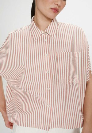 Femme portant une chemise ample à manches courtes avec des rayures verticales rouges et blanches, une poche avant et des boutons blancs, debout devant un fond uni.