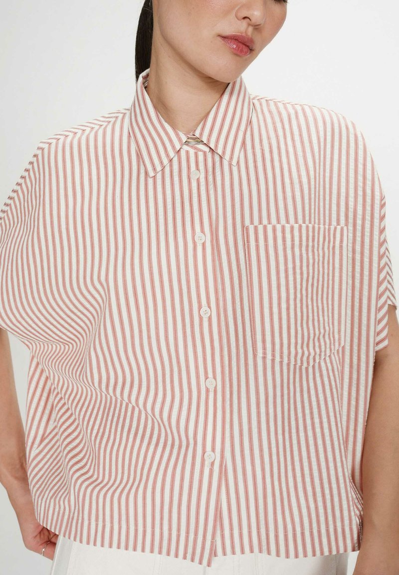 Mujer con una camisa holgada de manga corta con rayas verticales rojas y blancas, bolsillo delantero y botones blancos, de pie contra un fondo liso.