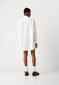 Hvit kjole med lange ermer, rett snitt, brystlappkrage og litt oversized passform, kombinert med svarte sko og hvite sokker.