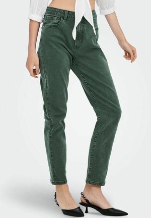 Person iført højtaljede grønne jeans, en hvid cropped bluse og sorte spidse hæle med kædedetalje, stående mod en ensfarvet baggrund.