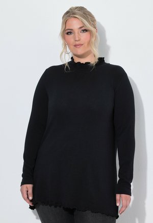 Blond vrouw in een zwarte top met lange mouwen en ruches aan de hals en zoom, staat tegen een witte achtergrond, kijkt naar voren.