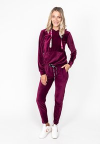 Violetti samettinen verkkapuku, jossa on huppu, etutasku ja säädettävä nyöri. Yhdistetty valkoisiin tennareihin, joissa on ruskea aksentti.