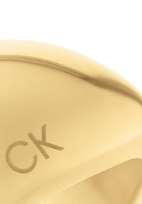 Złoty metalowy pierścień o gładkiej, zakrzywionej powierzchni; z wytłoczonym logo "CK"; błyszczące wykończenie z subtelnymi refleksami.