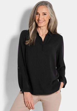 Schwarze Bluse mit V-Ausschnitt, langen Ärmeln und geknöpften Manschetten. Glatte Textur und taillierte Passform. Getragen mit hellen Hosen.