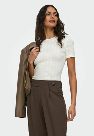 Femme aux longs cheveux foncés portant un haut blanc à manches courtes et un pantalon marron taille haute, tenant une veste marron sur son épaule.