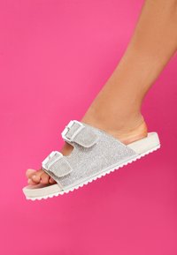 Sølvglitter slide sandaler med to justerbare hvide spænder og en struktureret fodseng, præsenteret mod en lyserød baggrund.