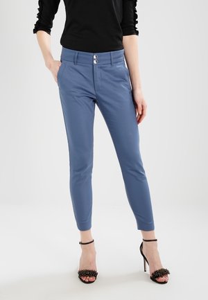 Kobieta ubrana w dopasowane niebieskie spodnie do kostek z podwójnym guzikiem w pasie oraz czarne sandały na wysokim obcasie z paskami wokół kostki.