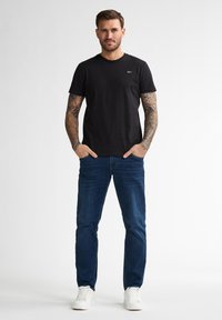 Camiseta de algodón negra con cuello redondo, combinada con jeans de mezclilla azules y zapatillas blancas. El modelo tiene tatuajes en ambos brazos.