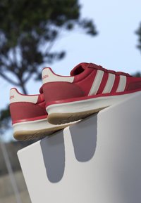 Baskets rouges Adidas avec des rayures blanches et des semelles en gomme reposant sur une surface blanche avec des arbres flous et un ciel bleu en arrière-plan.
