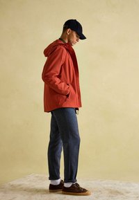 Vīrietis sarkanu kapuci jaku, tumši zilu cepuri, pelēkas bikses, baltas zeķes un brūnus sporta apavus stāv uz gaiša paklāja pret krēmkrāsas sienu.