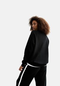 Sudadera negra de corte relajado, con cuello redondo y puños acanalados, combinada con pantalones negros que presentan rayas blancas en los laterales. Textura suave.
