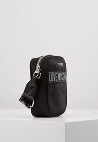 Černá křížová taška z tkaniny se stříbrným zipem, nastavitelným popruhem a výrazným logem "LOVE MOSCHINO" v šedé barvě na přední straně.