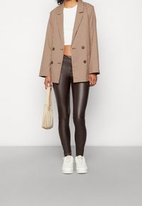 Brązowa marynarka w wzór houndstooth, biały crop top, błyszczące brązowe legginsy i białe sneakersy. Akcesorium: torebka na ramię w kolorze kremowym.