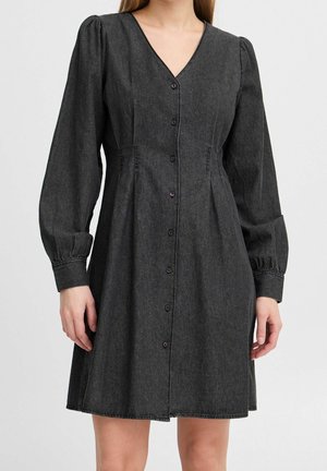 Frau trägt schwarzes Kleid mit Knopfleiste, langen Puffärmeln und V-Ausschnitt, steht vor einem schlichten hellen Hintergrund.