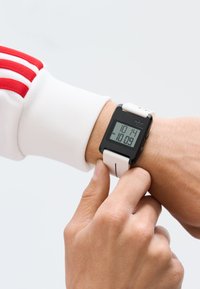 Digital klocka med svart urtavla, vit silikonrem, rektangulär form och LED-display som visar tiden. En hand justerar klockan på handleden.