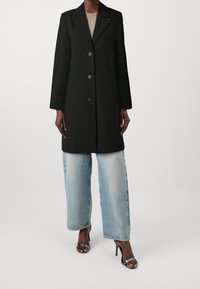 Abrigo largo tipo trench negro con cuello de muesca, botones delanteros abotonados y bolsillos laterales, combinado con jeans de pierna ancha en azul claro.