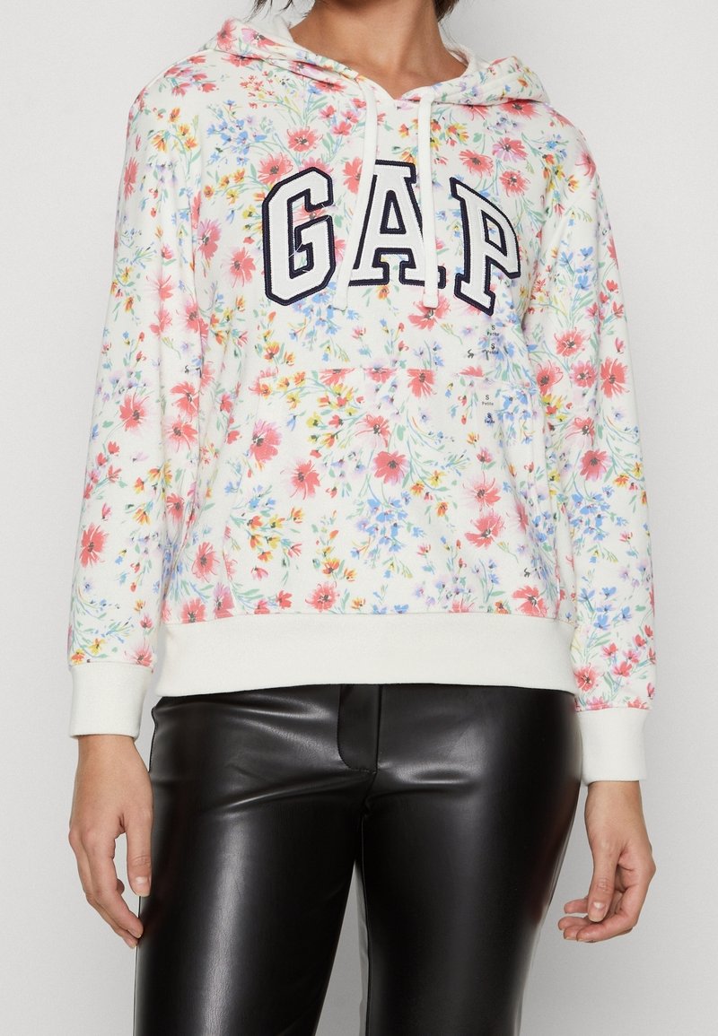 Femme portant un sweat à capuche blanc avec un imprimé floral coloré et le logo GAP noir, associé à un pantalon en cuir noir.