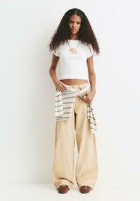 Kvinde med langt, krøllet hår iført en hvid crop top med hundemotiv, beige brede bukser, brune sko og en stribet sweater bundet om taljen.