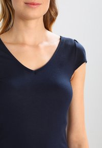 Námořnická modrá přiléhavá trička vyrobená z hladké tkaniny s V-výstřihem a krátkými rukávy, které mají jemné nabírané detaily.
