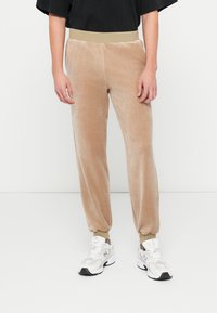 Pantalones de chándal de terciopelo beige con cinturilla y puños acanalados, que cuentan con una textura suave y un diseño ajustado, combinados con zapatillas blancas.