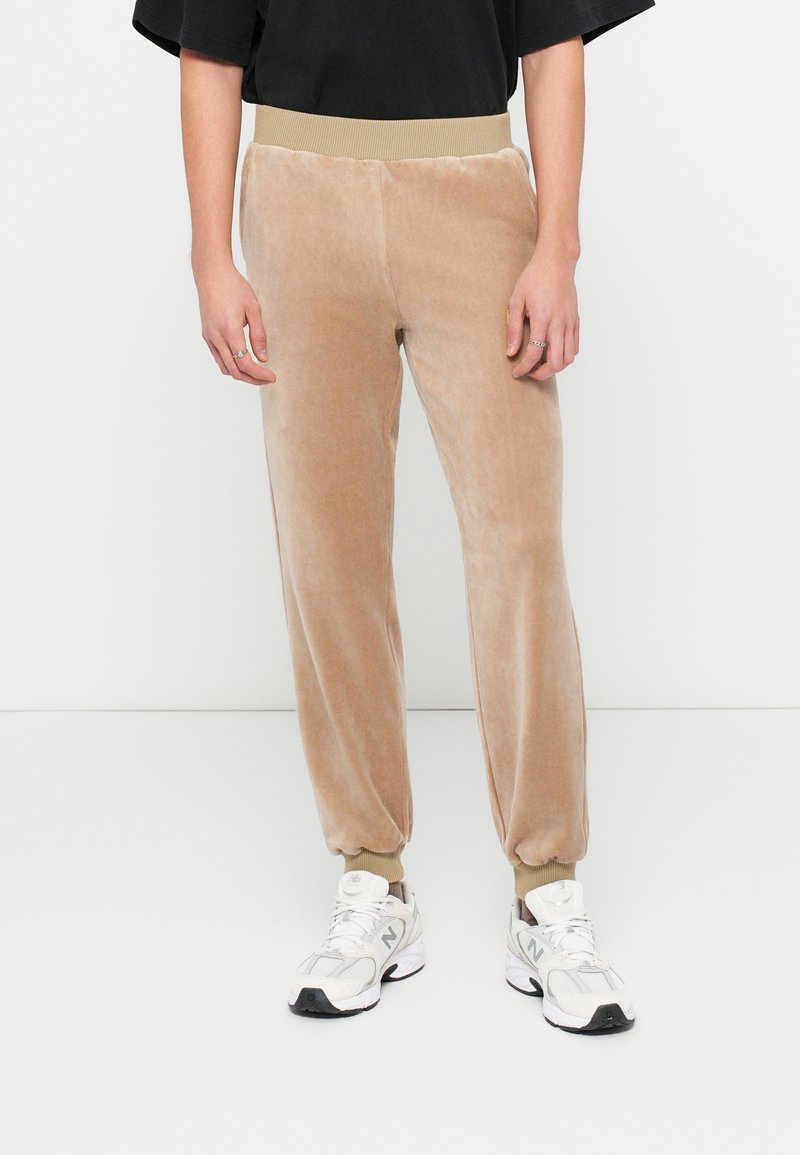Pantalones de chándal de terciopelo beige con cinturilla y pu�ños acanalados, que cuentan con una textura suave y un diseño ajustado, combinados con zapatillas blancas.