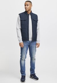 Marineblaue, gepolsterte Weste mit Reißverschluss, zwei Brusttaschen, getragen über einem grauen Sweatshirt und kombiniert mit hellblauen Jeans und marineblauen Sneakers.