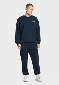 Granatowy sweter i dopasowane dresy wykonane z miękkiego materiału. Posiada okrągły dekolt i ściągacze przy mankietach. Nosi się z białymi sneakersami.