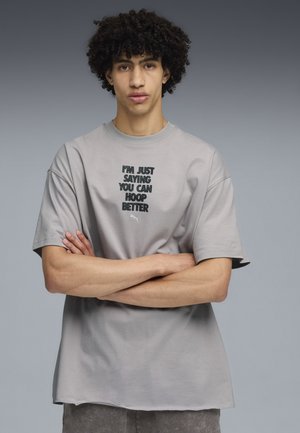 Graues Baumwoll-T-Shirt mit kurzen Ärmeln, featuring ein schwarzes Textdesign: "ICH SAGE NUR, DASS DU BESSER KORB SPIELEN KANNST" und ein kleines Logo.