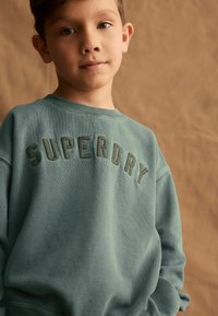 Tyrkysový sweatshir z bavlny s nápisom "SUPERDRY" vyšitým tmavozelenou farbou cez hruď. Mäkká textúra a dizajn s okrúhlym výstrihom.