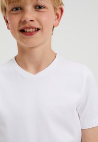 Biela bavlnená tričko s V-výstrihom a krátkymi rukávmi, s jednoduchým dizajnom a jemnou textúrou, nosené dieťaťom so svetlými blond vlasmi.