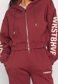 Burgundy cropattu huppari pehmeästä materiaalista, etuvetoketjulla, nyöreillä varustetulla hupulla ja valkoisilla logodetaileilla hihoissa ja vyötäröllä.