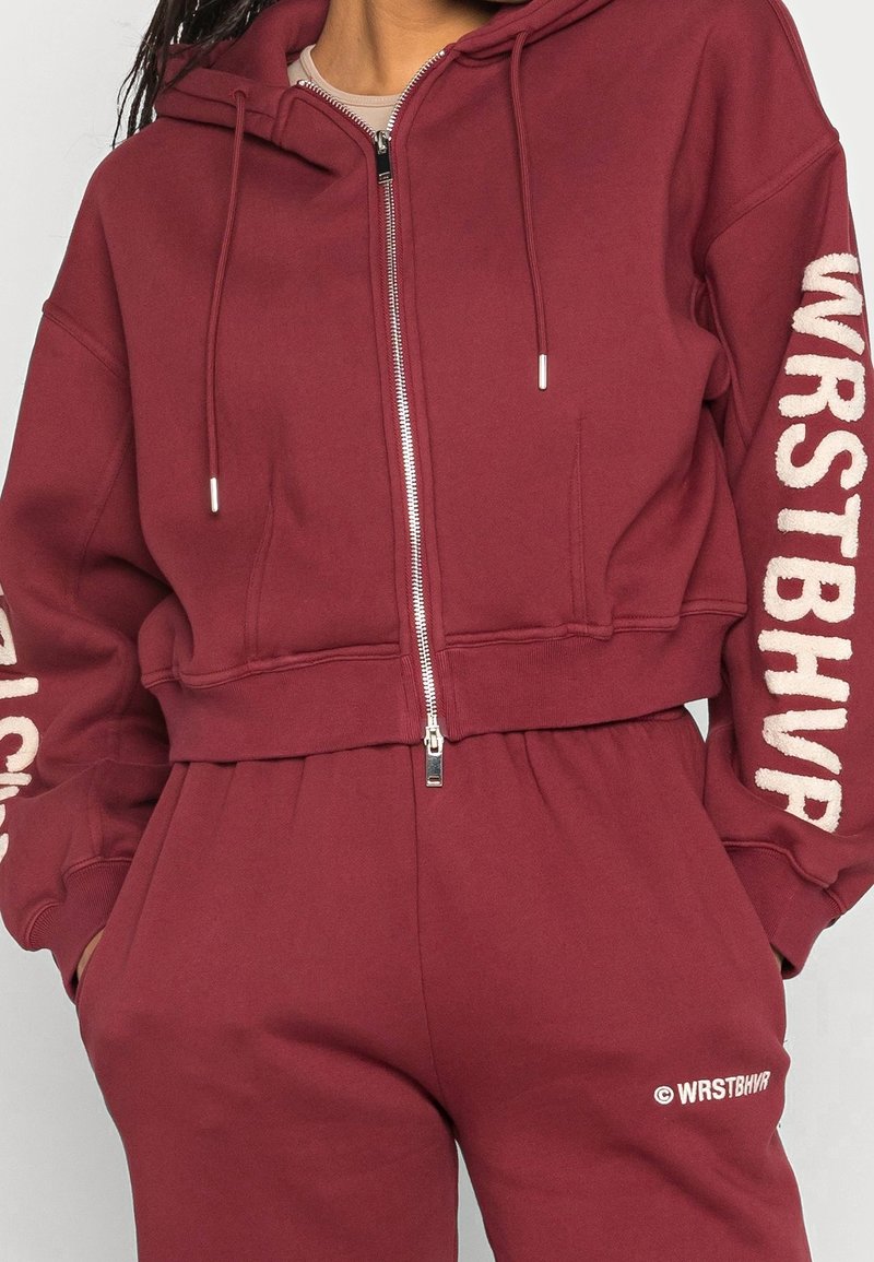 Burgundy cropattu huppari pehmeästä materiaalista, etuvetoketjulla, nyöreillä varustetulla hupulla ja valkoisilla logodetaileilla hihoissa ja vyötäröllä.