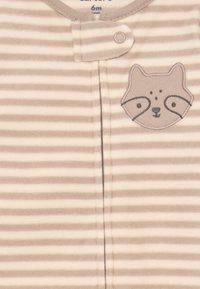 Stribet, blødt fleece-heldragt i beige og creme, med et broderet vaskebjørn-ansigt på brystet og trykknaplukning.