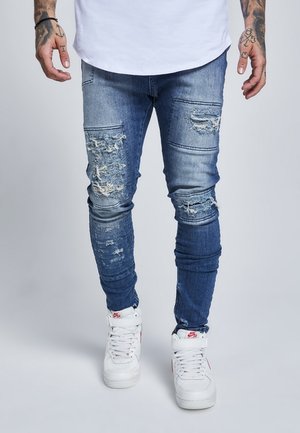 Mann trägt zerrissene blaue Skinny-Jeans, weiße hohe Nike-Sneaker und ein weißes Hemd, steht mit sichtbaren tätowierten Armen.