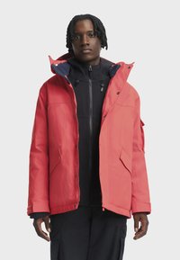 Jaunas vyras su trumpais dredais, vilkintis ryškiai raudoną žieminį švarką virš juodos užtrauktuku užsegamos maikutės ir juodų kelnių, stovi prieš pilką neutralų foną.