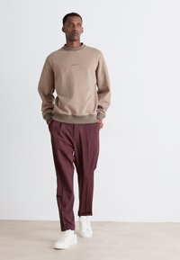 Tan sweatshirt with a textured finish and a subtle logo, paired with maroon trousers. Model wears white sneakers, standing on a light background.