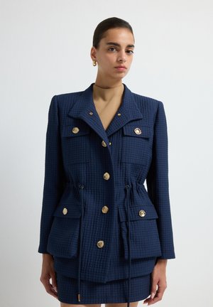 Blazer texturé marine avec des boutons dorés, comprenant deux poches poitrine et un cordon de serrage réglable à la taille. Associé à une jupe coordonnée.