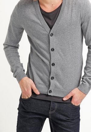 Mann trägt graue, geknöpfte Strickjacke über dunklem T-Shirt und dunklen Jeans, die Hände in den Hosentaschen.