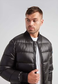 Giacca a vento nera con chiusura a zip e polsini a coste. Presenta una texture lucida e una trapuntatura orizzontale. Indossata sopra una maglietta bianca.