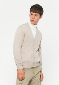 Beige strikket cardigan med V-hals, knappelukking og lommer. Har ribbestrikkede mansjetter og nederkant, kombinert med en hvit høyhalset genser.